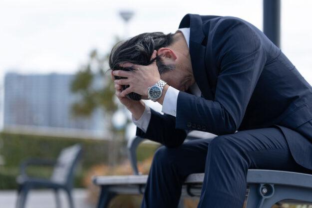 a person in a suit sits on an outdoor bench with their head in their hands while dealing with the long term effects of anxiety